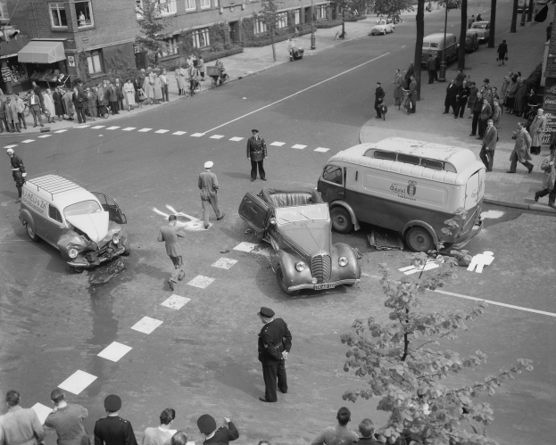 Schwarz-weiß-Szene eines Autounfalls an einer Straßenkreuzung mit mehreren Fahrzeugen, darunter ein Van, und einer Gruppe von Schaulustigen; Gebäude, Bäume, Strommasten und Schilder sind im Hintergrund zu sehen.