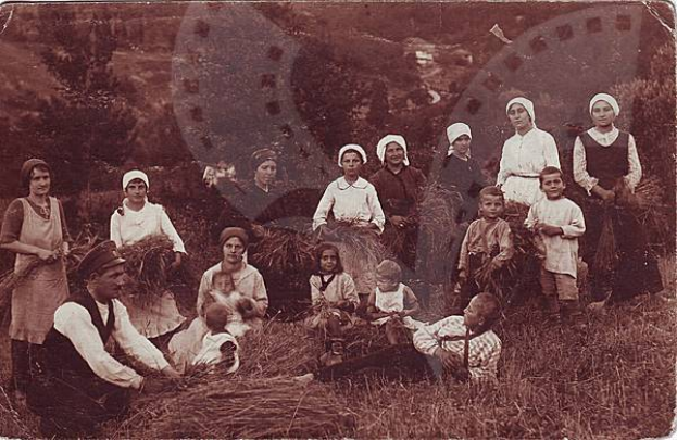 Ein altes Schwarz-Weiß-Foto einer Gruppe von Menschen in einem ländlichen Feld, einige stehen und einige sitzen auf dem Gras, mit Bäumen und einem Rad im Hintergrund, die traditionelle Kleidung tragen und verschiedene Werkzeuge und Geräte halten.