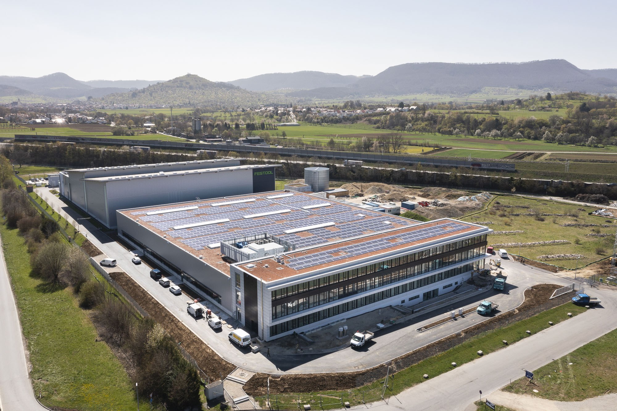 Luftaufnahme eines großen Gebäudes mit Solarmodulen auf dem Dach, umgeben von Fahrzeugen, Bäumen, Gras und Masten, mit Hügeln und einem klaren blauen Himmel im Hintergrund; das Gebäude ist die erste solarbetriebene Fabrik Deutschlands.