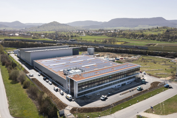 Luftaufnahme eines großen Gebäudes mit Solarmodulen auf dem Dach, umgeben von Fahrzeugen, Bäumen, Gras und Masten, mit Hügeln und einem klaren blauen Himmel im Hintergrund; das Gebäude ist die erste solarbetriebene Fabrik Deutschlands.
