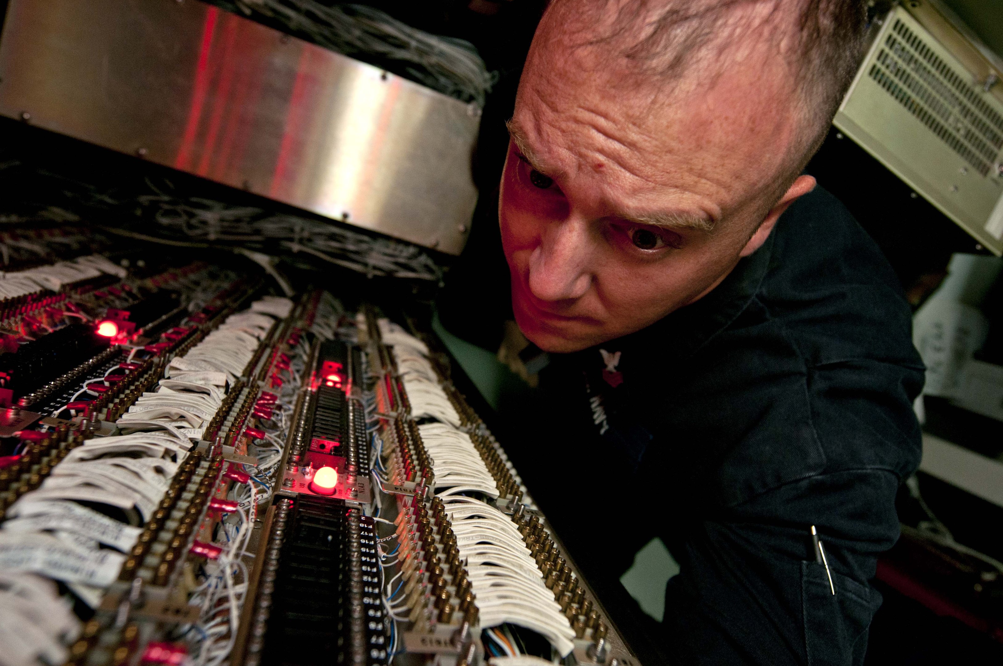 Ein Mann in einem schwarzen Hemd arbeitet an einem Computer und ist von elektronischen Geräten und Kabeln umgeben.