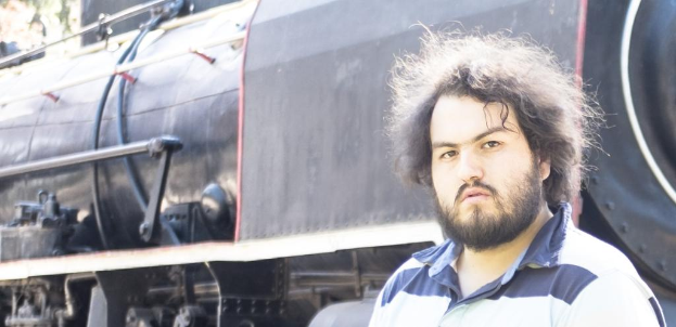 Ein Mann in einem T-Shirt steht neben einem Zug, wobei das Zugrad und andere Teile unten sowie ein Baum oben links zu sehen sind.