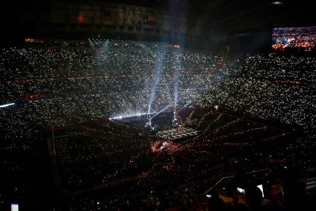 Eine große Menschenmenge in einem hell erleuchteten Stadion bei Nacht schaut auf einen Bildschirm mit den Ergebnissen der Super Bowl XLVII-Halbzeitshow.