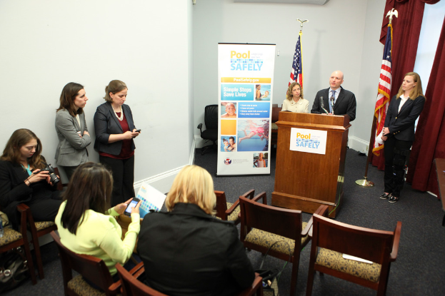 Drei Frauen sitzen auf Stühlen, während ein Mann und eine Frau vor einem Rednerpult stehen, mit zwei Flaggen und einem Banner im Hintergrund; links sieht eine Frau auf eine digitale Kamera und zwei andere Frauen stehen, und Vorhänge sind oben rechts im Bild zu sehen.