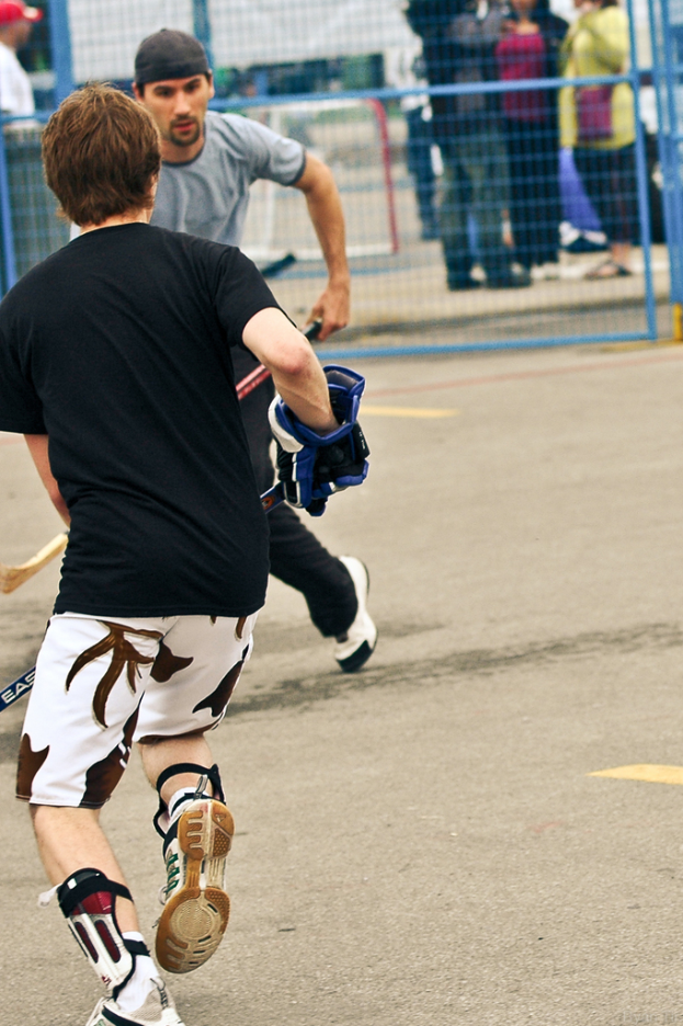 Zwei Personen spielen Hockey auf einer Straße mit einem Zaun dahinter und einer Menge Menschen im Hintergrund.