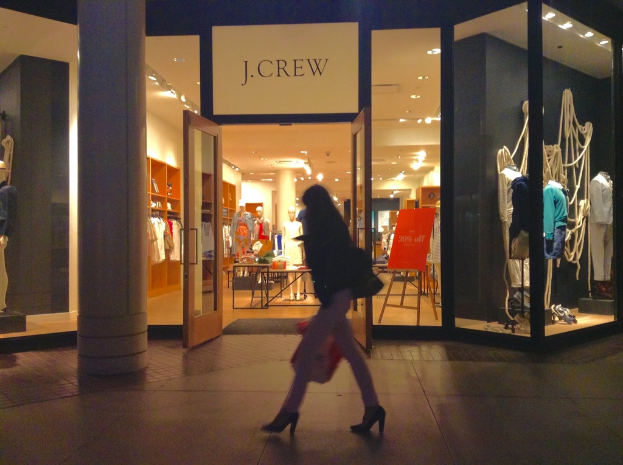 Ein Einkaufskomplex mit Glaswänden und -türen, der Puppen, Tische, hängende Kleider und Schränke zeigt, mit einer Frau daneben.