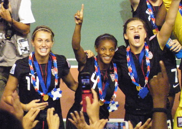 Das US-amerikanische Frauenfußballteam steht in schwarzen Kleidern und Medaillen auf einem Feld und feiert ihren 2014er WM-Sieg mit einem Fotografen und Zuschauern im Hintergrund.