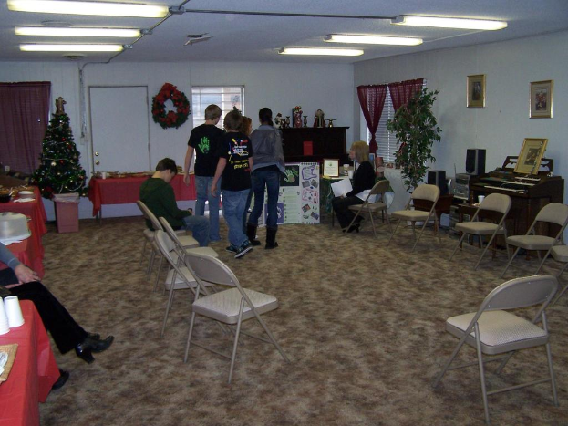 Ein Raum mit mehreren Stühlen, Tischen und stehenden und sitzenden Personen, mit einem Weihnachtsbaum, einem Piano, einer Wand mit einem Strauß, Vorhang, Tür, Fotorahmen, Fenstern und Deckenröhren.