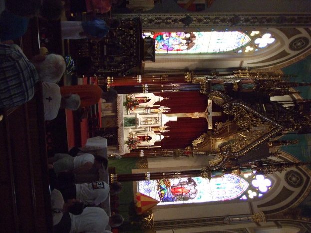 Ein Innenblick in eine Kirche mit einem Kruzifix, das eine Jesus-Skulptur zeigt, und zahlreichen Menschen, die vor ihm auf Stühlen sitzen.