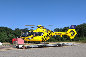 Ein gelber Hubschrauber auf einem Flughafenvorfeld mit verschiedenen Gegenständen drumherum, umgeben von Bäumen unter einem klaren blauen Himmel, wahrscheinlich für Luftrettungsdienste in Deutschland verwendet.