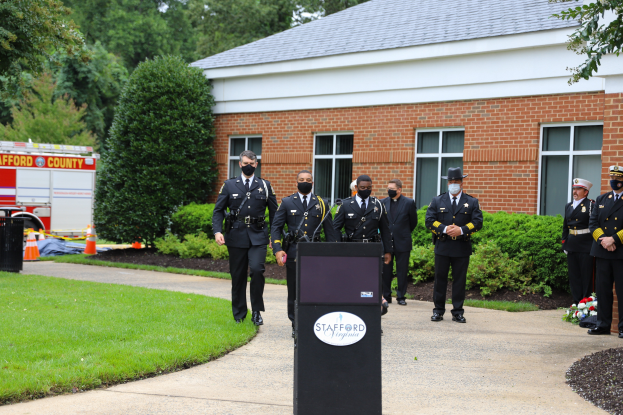 Eine Gruppe von Polizisten in Uniformen und Masken steht vor einem Gebäude mit Fenstern und Türen, mit einem Podium, Mikrofon und Texttafel im Vordergrund, Gras links und einem Löschfahrzeug, Bäumen, Pflanzen, Blumen, Verkehrskegeln und anderen Gegenständen im Hintergrund unter einem sichtbaren Himmel.