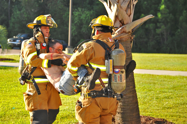 Drei Feuerwehrleute tragen einen vierten Feuerwehrmann auf einer Trage über eine grüne Fläche mit Bäumen, Fahrzeugen und einem Mast im Hintergrund.