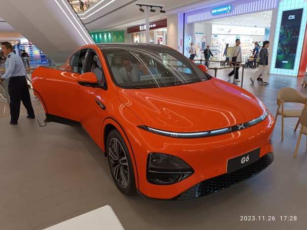 Oranges Elektroauto in einer Ausstellungshalle mit einer Gruppe von Menschen drumherum, Stühlen und einem Wagen rechts, Geschäften und Beschilderung im Hintergrund.