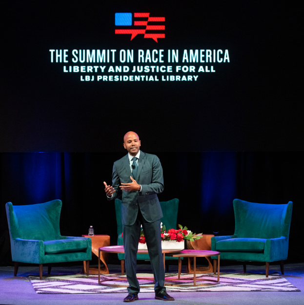 Ein Mann in einem Blazer, Krawatte und Hemd steht auf einer Bühne vor zwei Stühlen und einem Tisch mit einem Blumenstrauß und einer Flasche, mit einem Vorhang und einem Bildschirm, der "The Summit on Race in America: Liberty and Justice for All" anzeigt, dahinter.