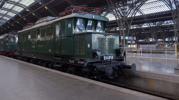 Ein Zug auf Eisenbahnschienen mit Geländern und Tafeln im Hintergrund, Lampen auf dem Dach und einen Himmel darüber, mit einem Boden darunter.