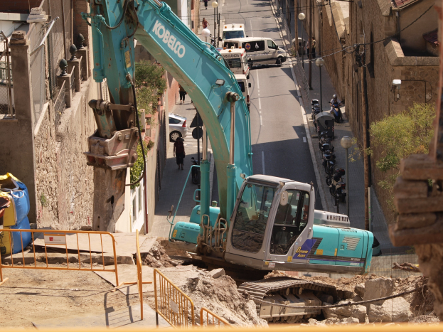 Bagger bei Bauarbeiten auf einer Stadtbaustelle mit Gebäuden, Straßeninfrastruktur, Fahrzeugen, Passanten und Bäumen im Hintergrund.