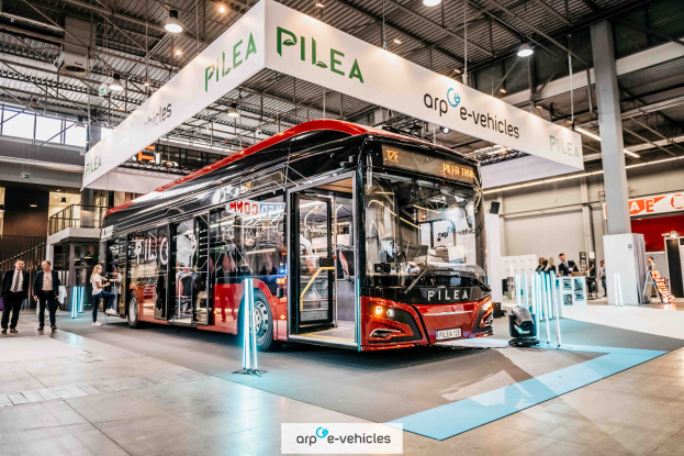 Ein roter Bus steht in einem Gebäude, umgeben von Menschen, mit einem Schild, auf dem "Pilea e-Fahrzeuge auf der Frankfurter Automobilausstellung" steht, Pfosten, eine Treppe, ein Geländer, eine Wand, ein Dach mit Metallpfosten und Deckenlampen.