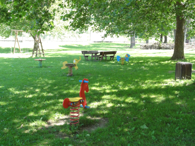 Eine Parkszene mit Schaukeln, Bänken und einem Baum auf grünem Rasen, umgeben von üppigen Bäumen.
