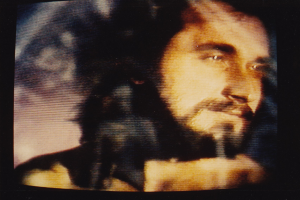 Ein Mann mit Bart, Schnurrbart und langen dunklen Haaren trägt ein weißes Hemd und schaut direkt in die Kamera auf einem TV-Bildschirm mit dunklem Hintergrund.