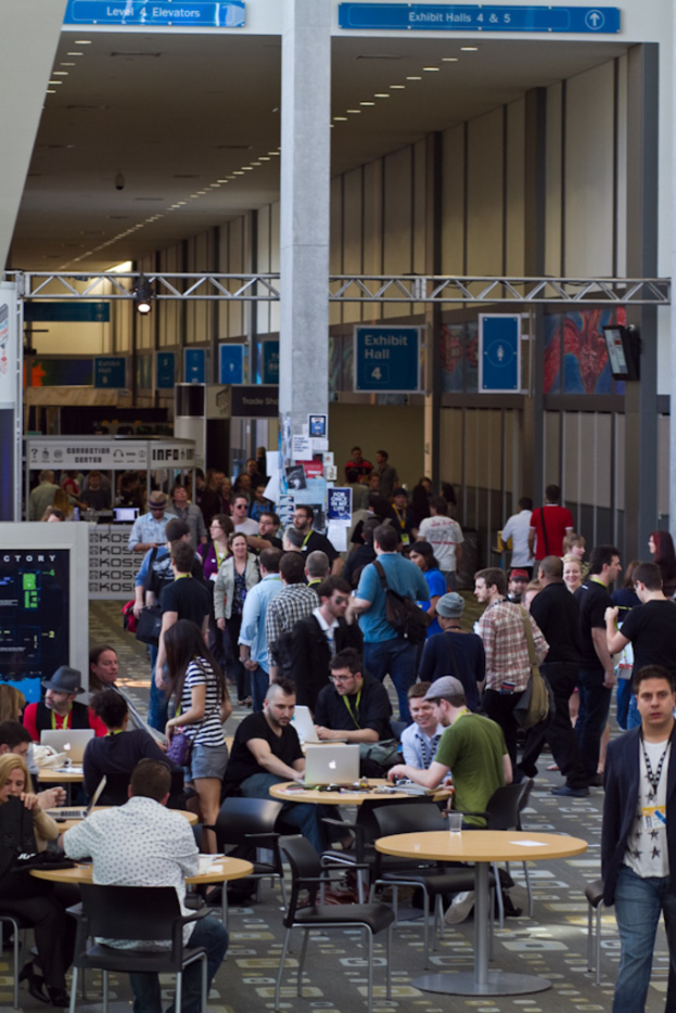 Ein belebter Flughafenvorplatz mit wartenden, arbeitenden und verschiedenen Aktivitäten nachgehenden Menschen, Richtungsschilder an der rechten Wand und darüber sowie Deckenbeleuchtungskörper.