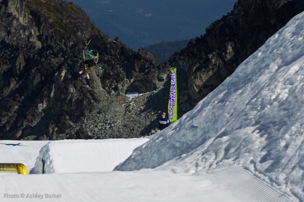 Ein Mann in Skier springt in die Luft mit schneebedeckten Bergen und einer Flagge mit Text im Hintergrund und einem stehenden Mann vor der Flagge, mit Text in der unteren linken Ecke.