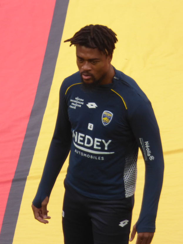 Ein Mann in einem blauen Hemd und schwarzen Hosen läuft auf einem Fußballfeld, mit einem Banner im Hintergrund, während eines Trainings.