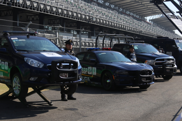 Eine Gruppe von Polizeiwagen vor einer Rennstrecke geparkt, uniformierte Personen auf der Straße stehend und im Hintergrund ein Stadion mit Geländern und Maschendrahtzaun sichtbar.