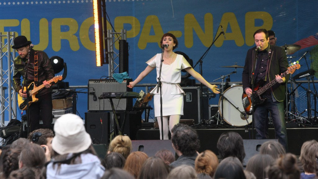 Vier Musiker auf der Bühne: eine Frau in einem weißen Kleid singt in der Mitte, flankiert von zwei Männern, die Gitarren spielen, mit einer Menge unten und Lautsprechern an der Wand dahinter.