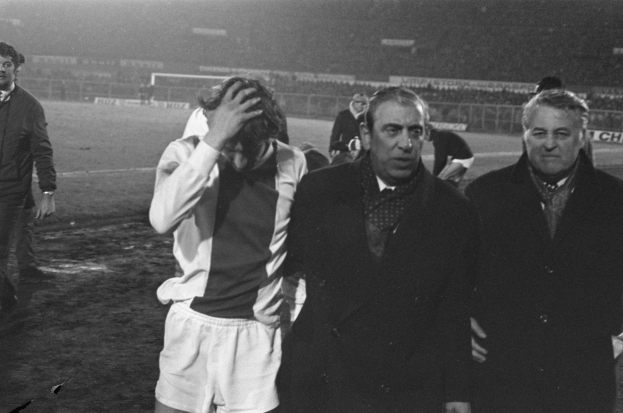 Eine Gruppe von Männern in Not steht zusammen auf einem Fußballfeld mit einem Stadion im Hintergrund, aufgenommen in Schwarz-Weiß.