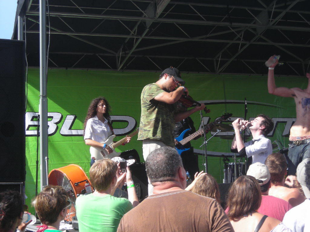 Eine Gruppe von Musikern spielt verschiedene Instrumente auf einer Bühne mit einem großen schwarzen Lautsprecher links daneben, während das Publikum vor einem Podium steht und die Vorstellung genießt, und ein grünes Banner im Hintergrund.