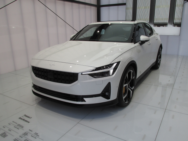 Ein weißer Polestar 1 Elektroauto in einer Ausstellungshalle, das eine schlankes, modernes Design mit einem glänzenden Finish, hellen, winkligen Scheinwerfern, einer langen Motorhaube, einem tiefgelegten Körper, großen Sporträdern und einem geräumigen, luxuriösen Innenraum mit Ledersitzen und einem modernen Armaturenbrett aufweist.