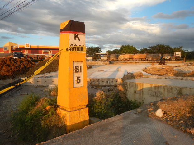Eine Baustelle mit einem gelben Warnschild auf der Straße, eine Säule mit Text und Zahlen im Vordergrund, Drähte links und Fahrzeuge, Bäume, ein Gebäude und der Himmel im Hintergrund.