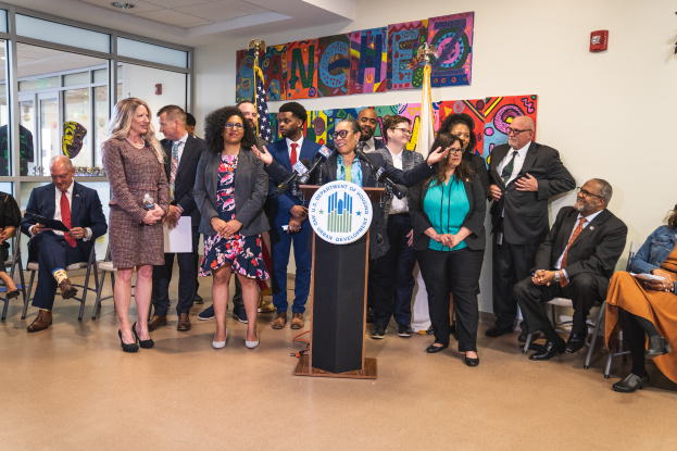 Eine Gruppe von Menschen, die vor einem Podium mit Mikrofon und einer Tafel mit der Aufschrift "Sancho-Stadtrat tritt dem Stadtrat bei" stehen und sitzen, mit Flaggen, Feuerlöscher und Deckenleuchten an der Wand im Hintergrund.