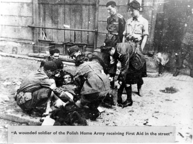 Ein verletzter Soldat der Polnischen Heimatarmee liegt auf dem Boden in einem Schwarz-Weiß-Bild, der erste Hilfe von einigen stehenden Individuen erhält, mit einer Wand und einem Geländer im Hintergrund.