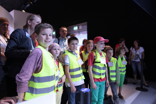Eine Gruppe von Kindern mit Sicherheitswesten und Namensschildern, wobei ein Junge auf der linken Seite lächelt, auf einem Boden mit einem Pfahl auf der rechten Seite und einigen Menschen im Hintergrund.