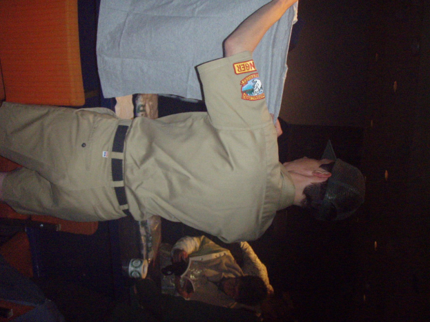 Ein Polizeibeamter hält ein T-Shirt in den Vordergrund, während ein Junge im Hintergrund ein Buch und einen Becher hält und in der Nähe eines Schreibtischs und Lampen steht.