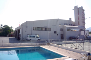 Ein großes Gebäude mit einem Schwimmbad davor, umgeben von einem Metallzaun, mit Fahrzeugen, Pfählen, Drähten, einem Straßenpfahl, Bäumen, Hügeln und einem bewölkten Himmel.