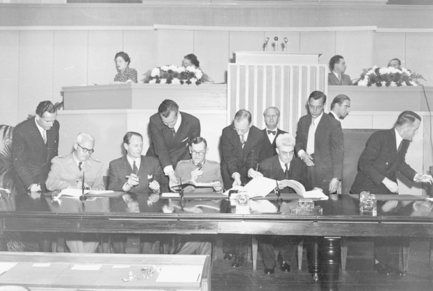 Eine Gruppe von Menschen sitzt auf Stühlen mit anderen dahinter an einem Tisch mit Mikrofonen, Gläsern und Blumenbouquets.