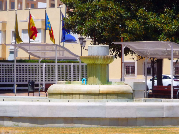 Ein Brunnen in einem park├Ąhnlichen Gebiet mit einem umzünten Geb├Ąude, Flaggen, einem Schuppen, B├Ąnken, einem geparkten Auto, Laternenmasten und B├Ąumen im Hintergrund.