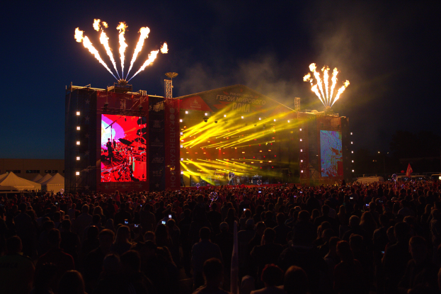 Eine große Menschenmenge versammelt sich auf einem Musikfestival, mit einer Bühne in der Mitte, auf der Musiker von hellen Scheinwerfern beleuchtet werden und von Bannern, Zelten links und einem dunklen Himmel mit Feuerwerk im Hintergrund umgeben sind.