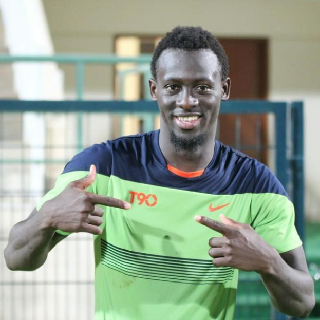 Ein Mann in einem grünen und blauen Hemd zeigt mit einem Lächeln auf die Kamera, im Hintergrund ist ein leicht unscharfer Geländer, eine Wand und eine Tür zu sehen.