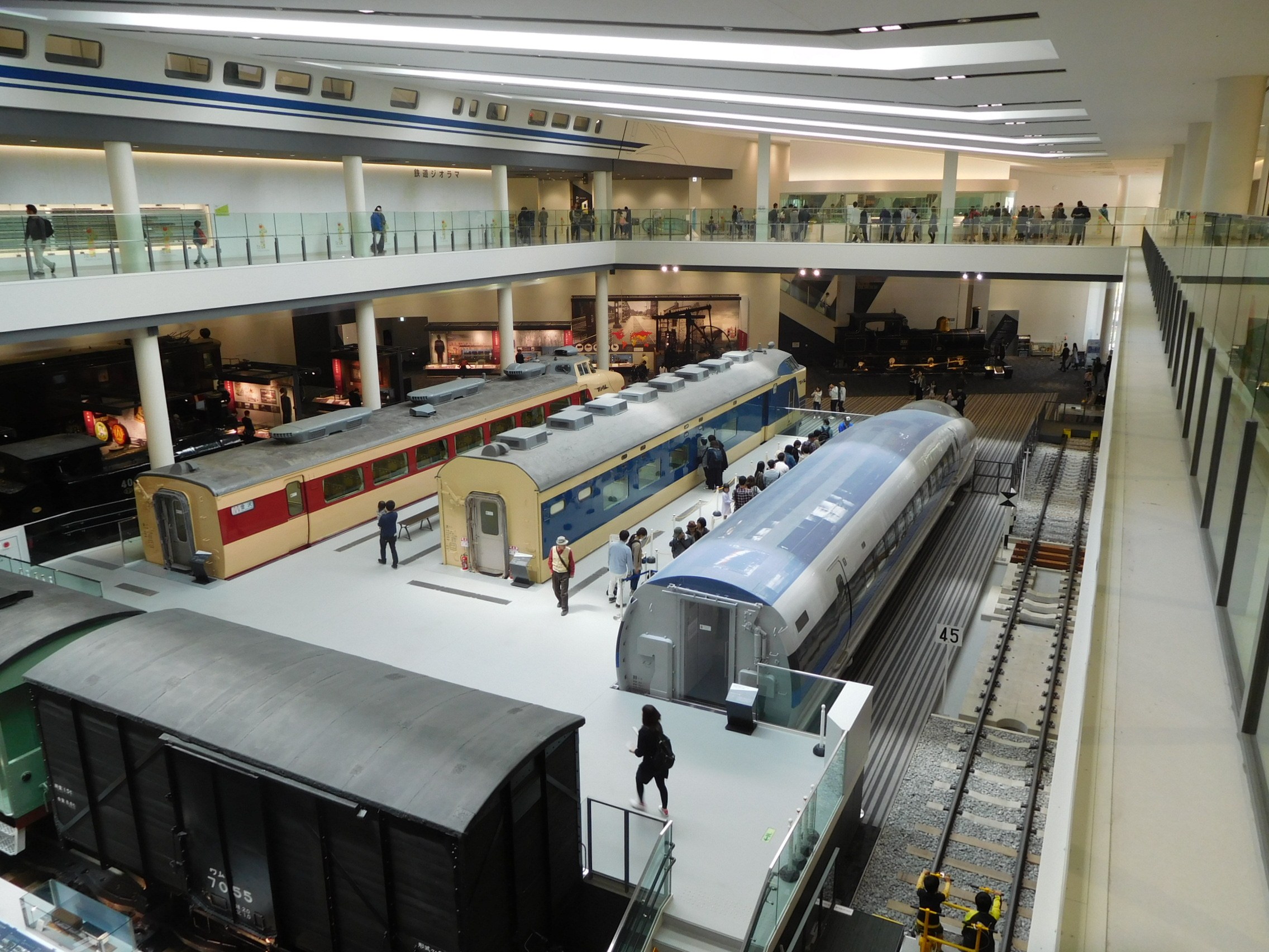 Ein großer Museumsraum mit verschiedenen Zügen in unterschiedlichen Größen und Farben, bei dem Menschen die Ausstellungsstücke und architektonische Elemente wie Geländer, Säulen und Deckenleuchten betrachten.