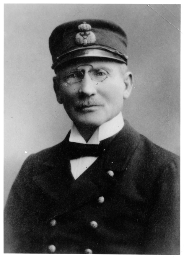 Ein Schwarz-Weiß-Foto eines Mannes in einer Mütze und mit Brille, der einen strengen Ausdruck hat und wohl ein Captain ist.