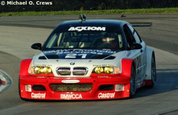 Eine Person in einem Helm fährt ein weißes BMW M3 GT3 Rennauto auf einer von Gras und einem Maschendrahtzaun umgebenen Strecke, mit sichtbarem Text und Zahlen auf dem Auto und einem Wasserzeichen in der oberen linken Ecke.