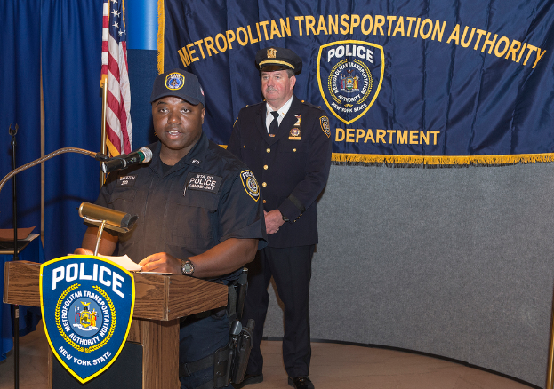 Zwei Polizeibeamte stehen vor einem Podium, einer hält Papiere und spricht in ein Mikrofon, mit einer Tafel, einer Fahne und einem Tisch in der Nähe und einer Tafel mit Symbolen und Text im Hintergrund.