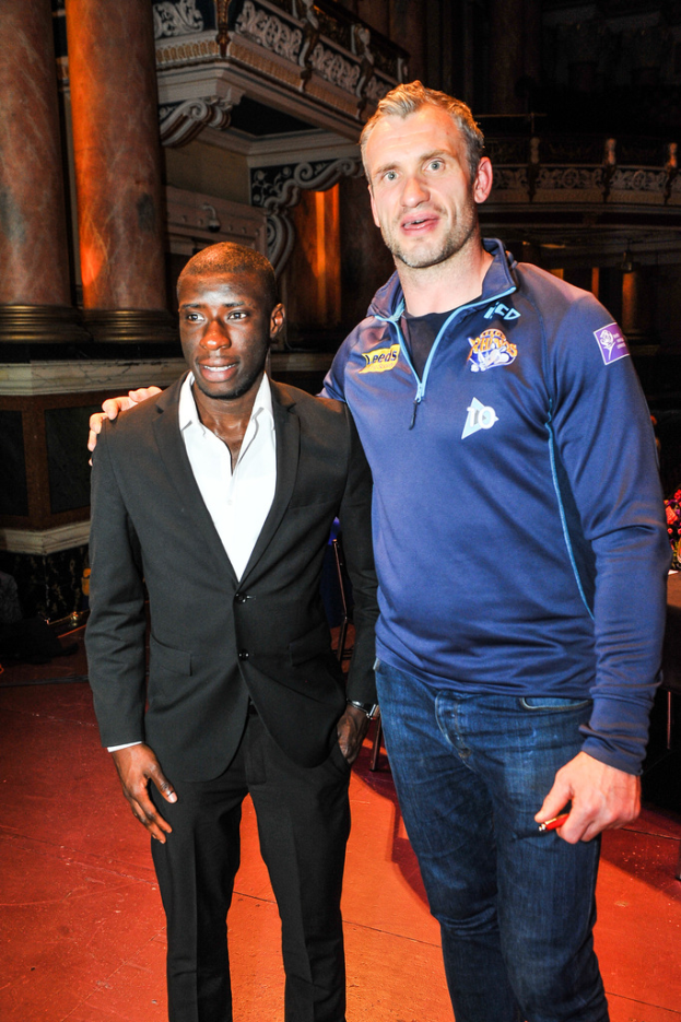 Zwei Männer stehen nebeneinander, wobei einer einen Anzug trägt und zwei Säulen im Hintergrund zu sehen sind.