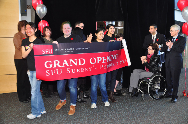Eine Gruppe von Frauen hält ein Banner, um ein Ereignis zu bewerben, mit zwei klatschenden Männern daneben und einer sitzenden, aufgeregten Frau im Rollstuhl vor einem schwarzen Vorhang und Ballons.