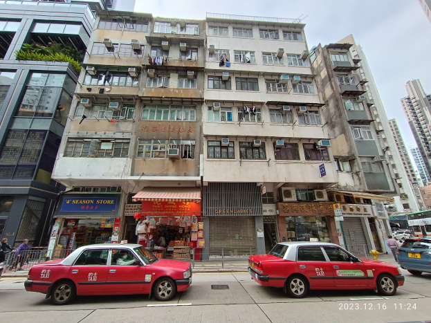 Rotes Taxi fährt auf einer Straße mit hohen Gebäuden, Passanten und verschiedenen städtischen Details unter einem bewölkten Himmel.