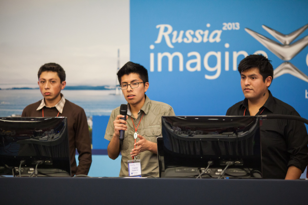 Drei Jungen stehen neben einem Schreibtisch mit zwei Computersystemen, einer spricht in ein Mikrofon und trägt eine ID-Karte mit roten Tags, vor einer Wand mit einer Werbetafel, auf der 'Russland Imagine 2013' steht.