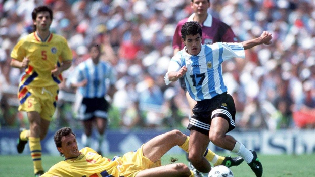Eine Gruppe von Fussballspielern auf einem Rasenfeld, mit Argentiniens Diego Maradona, der von Kolumbiens Diego Sanchez getackelt wird, und Zuschauern im Hintergrund.
